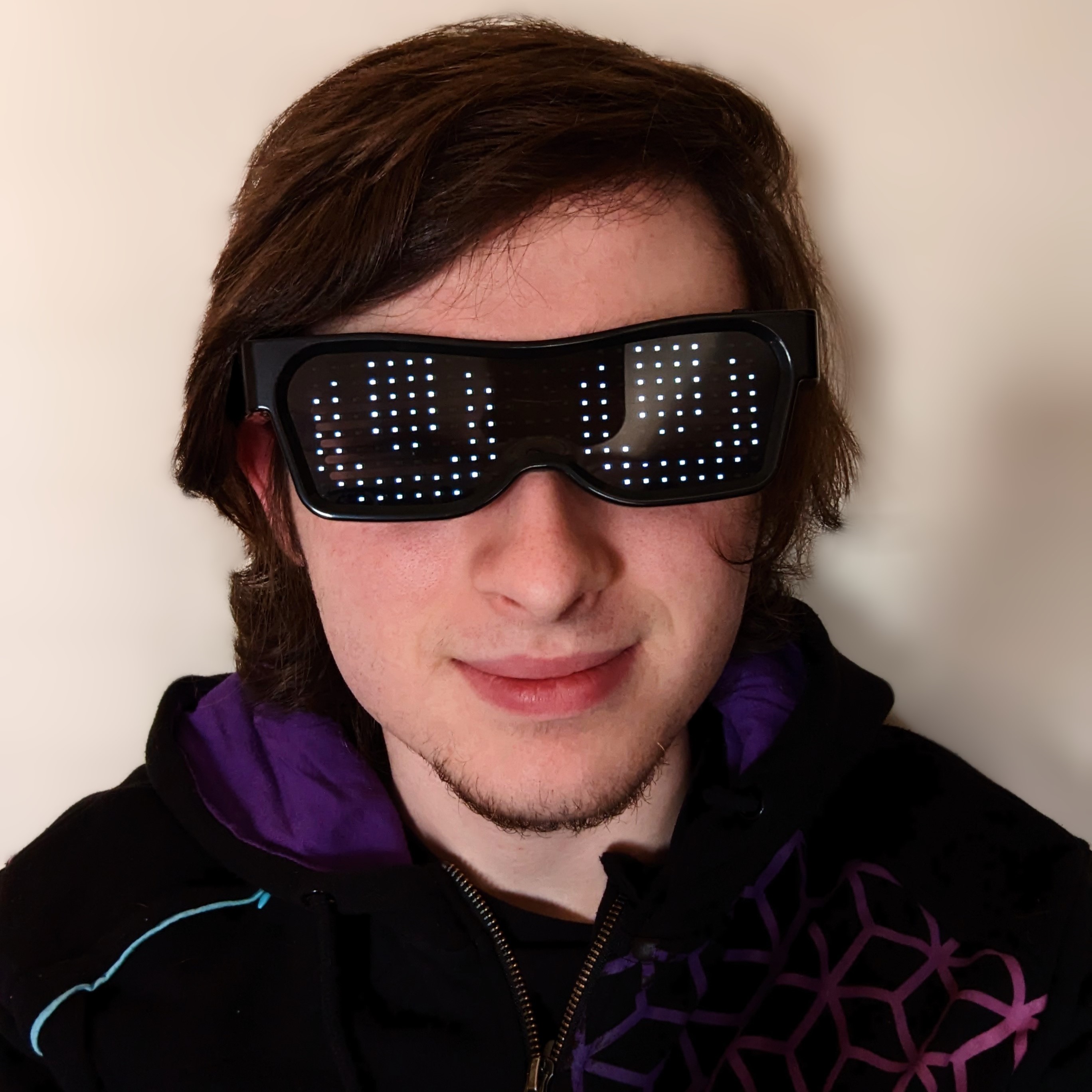 Brown-haired man wearing a black and purple jumper and LED glasses displaying robot eyes. The image is within a circular frame